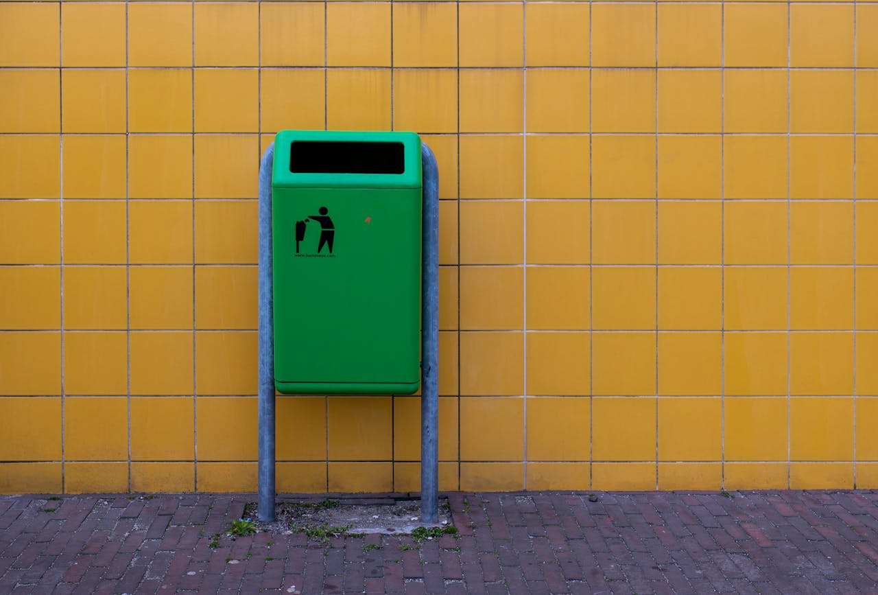A green trash bin standing against a vibrant yellow tiled wall, conveying urban cleanliness.